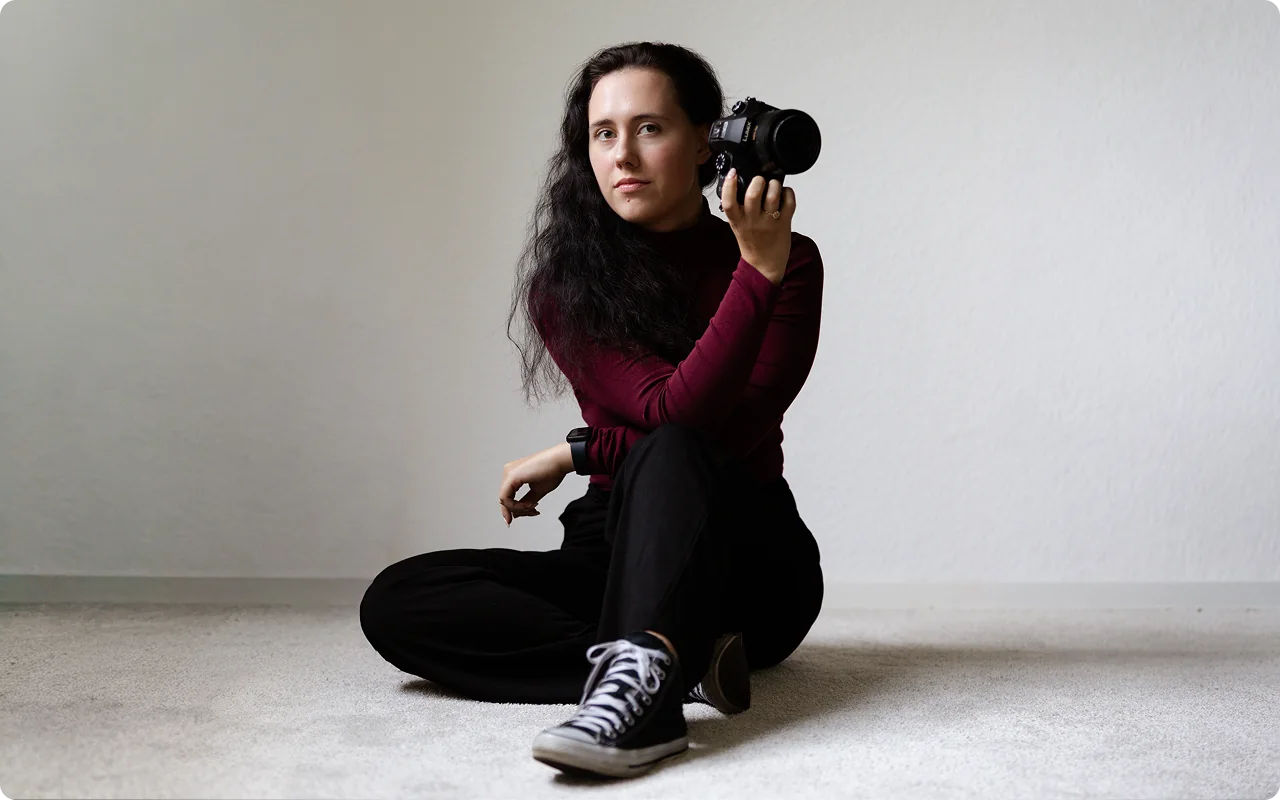 Die Fotografin sitzt im Schneidersitz auf einem hellen Teppich, hält ihre Kamera neben das Gesicht und blickt in die Kamera.