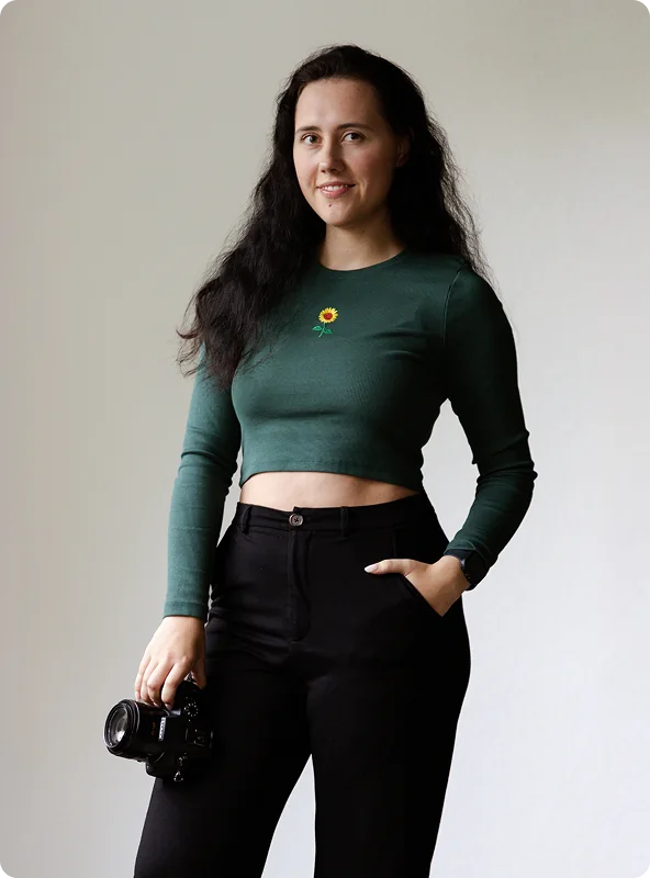 Die Fotografin steht in grünem Longsleeve mit kleiner Sonnenblume und schwarzer Hose, eine Hand in der Hosentasche, in der anderen ihre Kamera.