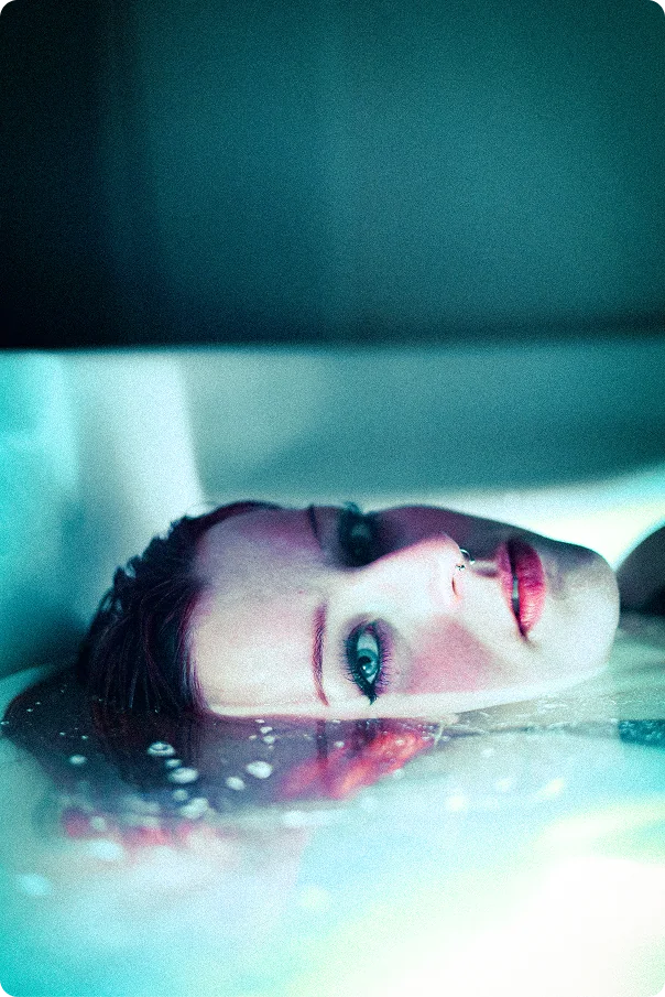 Das Gesicht einer Frau liegt halb unter Wasser, ihre hellen Augen blicken seitlich in die Kamera; im Wasser spiegeln sich rötliche Lichter.
