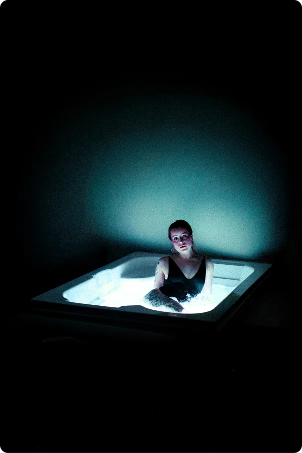 Weitwinkelaufnahme eines dunklen Raums, in dem eine beleuchtete Wanne leuchtet; darin sitzt eine kleine Gestalt mit Blick zur Kamera.