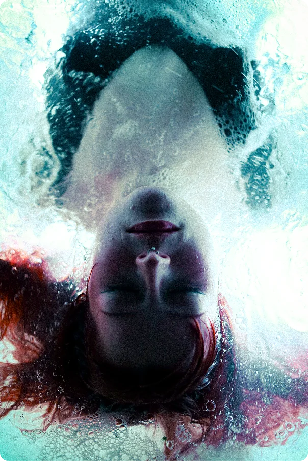 Unterwasseraufnahme von oben: Der auf dem Kopf stehende Kopf einer Frau mit geschlossenen Augen zwischen Luftblasen und leuchtendem Wasser.