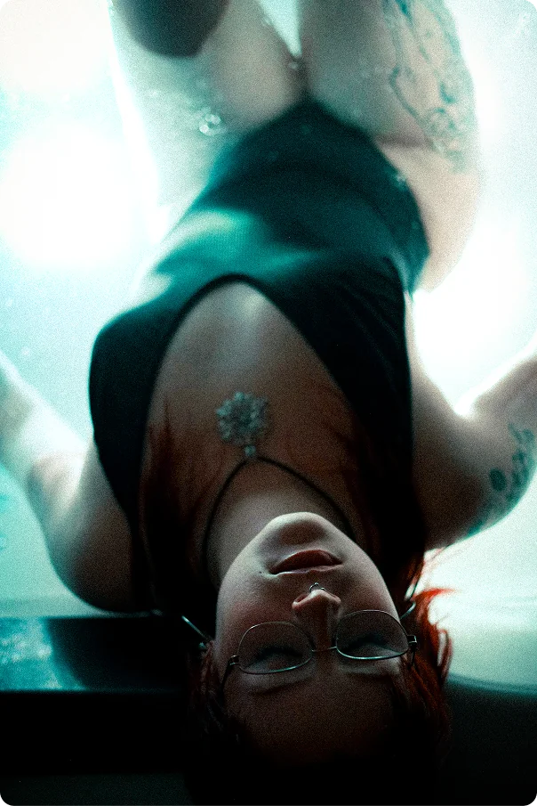 Auf dem Kopf stehende Aufnahme einer Frau mit Brille und roten Haaren im Wasser, der Blick geht nach oben, im Hintergrund leuchtet es türkis.