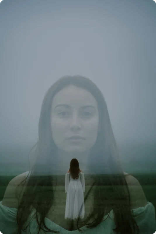 Doppelbelichtung im Nebel: Das riesige Gesicht einer Frau erscheint im Himmel, darunter steht eine kleine Gestalt in weißem Kleid mit dem Rücken zur Kamera auf einer Wiese.