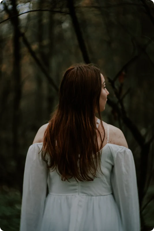 Eine Frau mit langen rotbraunen Haaren steht mit dem Rücken zur Kamera in einem dunklen Wald und blickt zur Seite; sie trägt ein weißes, schulterfreies Kleid.