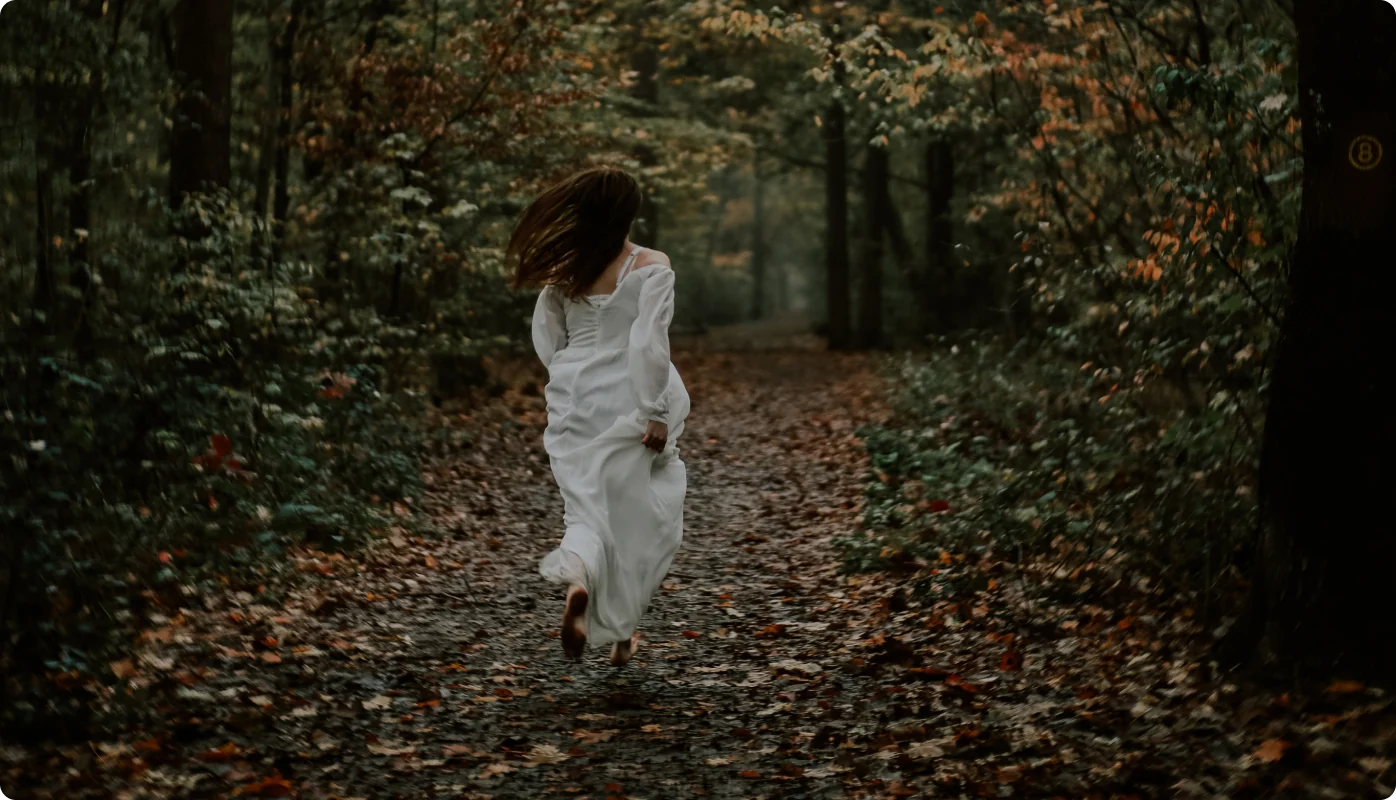 Eine Frau in weißem Kleid rennt barfuß mit wehenden Haaren einen von Herbstlaub bedeckten Waldweg entlang.