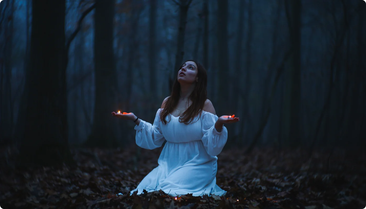 Eine Frau in weißem Kleid kniet in einem blau getönten, nebligen Wald und hält in beiden Handflächen kleine brennende Kerzen, den Blick nach oben gerichtet.