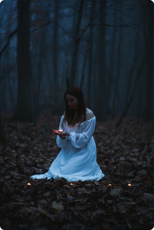 Eine Frau in weißem Kleid kniet zwischen Herbstlaub in einem blau getönten Wald und hält ein Teelicht in den Händen; vor ihr leuchten weitere kleine Kerzen.