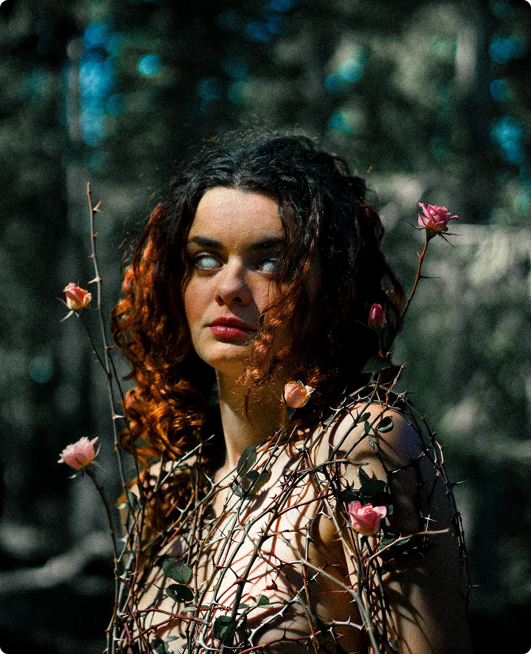 Experimentelles Porträt: Eine Frau mit dunklen Locken und hellen, fast weißen Augen; über ihre Schulter ranken sich dornige Rosenzweige mit rosa Blüten vor einem schattigen Wald.