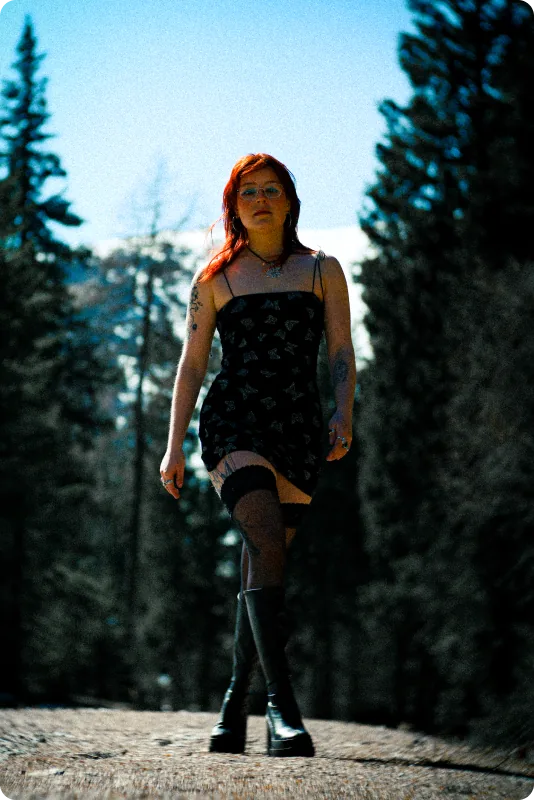 Eine Frau mit roten Haaren und Brille geht in schwarzem Kleid mit Schmetterlingsmuster, Strümpfen und Plateaustiefeln auf einem Waldweg direkt auf die Kamera zu.
