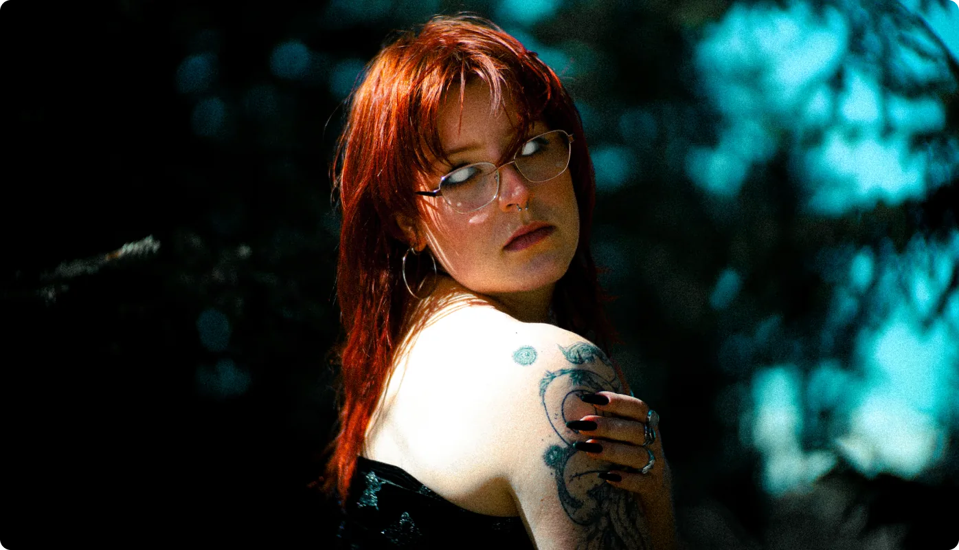 Eine Frau mit rotem Haar und Brille blickt über ihre tätowierte Schulter in die Kamera; um sie herum dunkler Wald mit türkisfarbenen Lichtreflexen.