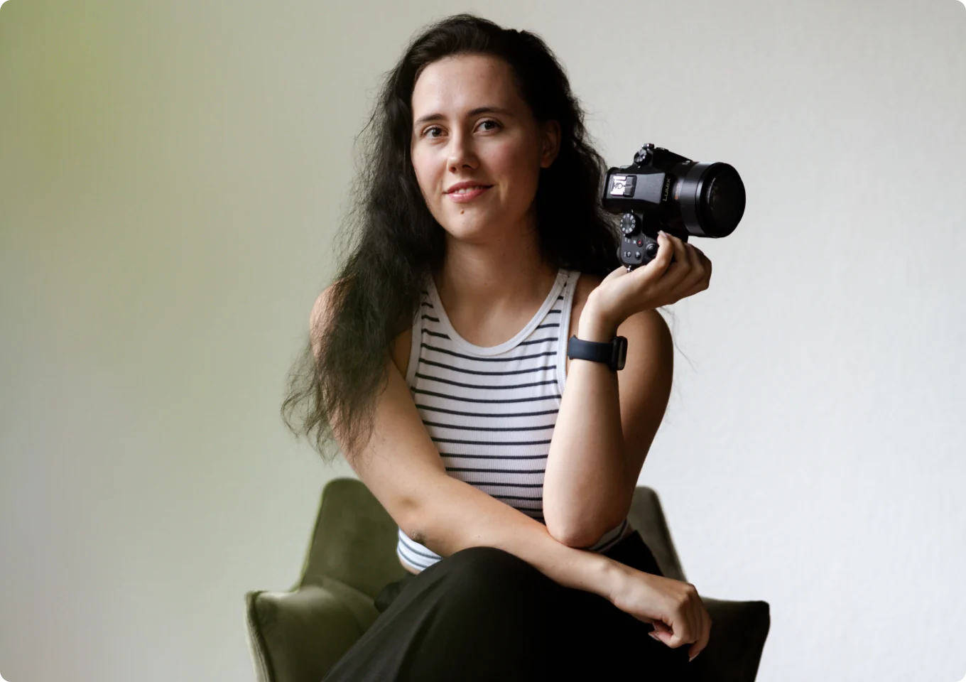 Porträt der Fotografin: Sie sitzt in gestreiftem Top auf einem olivgrünen Sessel, hält ihre Kamera in der Hand und blickt ruhig in die Kamera.