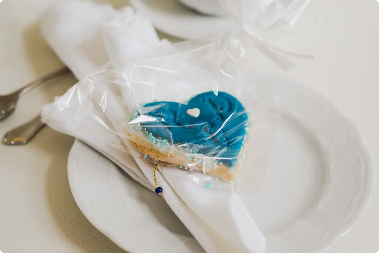 Ein Hochzeitsgebäck in Herzform mit blauem Zuckerguss liegt in Folie verpackt auf einer gefalteten weißen Serviette auf einem Teller.