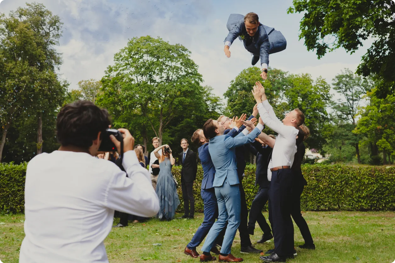 Der Bräutigam fliegt im Anzug hoch durch die Luft, unter ihm strecken Freunde die Arme aus; im Vordergrund fotografiert ein Gast, dahinter grüne Bäume.