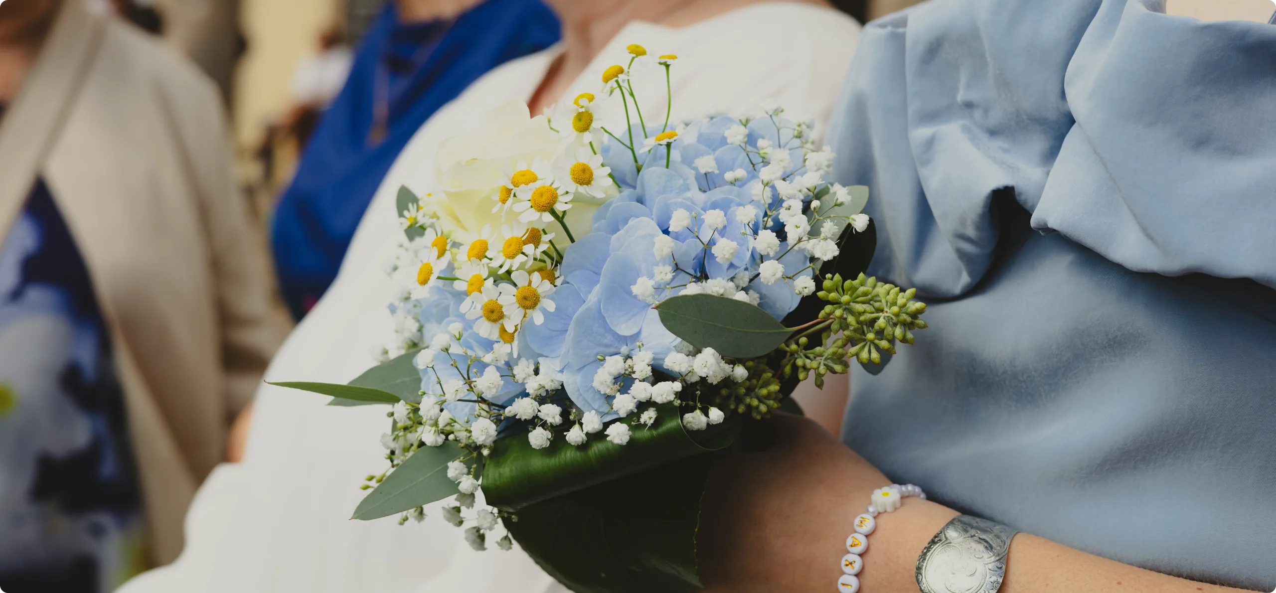 Ein Brautstrauß aus hellblauen Hortensien, weißer Rose, Kamille und Schleierkraut; daneben ein hellblaues Kleid mit Rüschen und ein Perlenarmband.