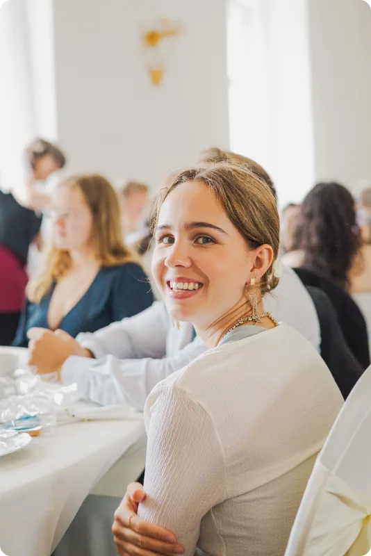 Eine junge Frau in hellem Oberteil sitzt an der festlich gedeckten Tafel und blickt lächelnd über die Schulter in die Kamera; im Hintergrund weitere Gäste.