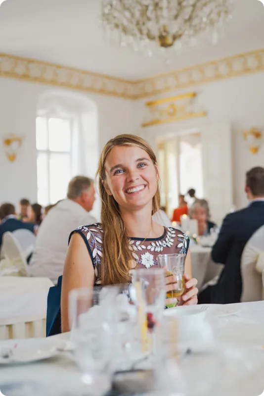 Eine junge Frau im Kleid mit Blumenstickerei sitzt lächelnd an der festlich gedeckten Tafel und hält ein Glas in der Hand; hinter ihr ein Saal mit Kronleuchter.