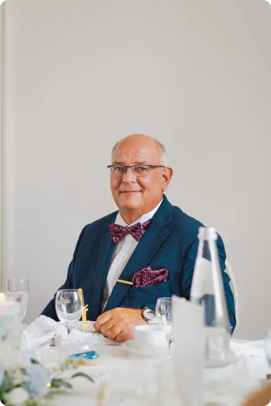 Ein älterer Herr im dunkelblauen Anzug mit Fliege und Brille sitzt lächelnd an der Hochzeitstafel.