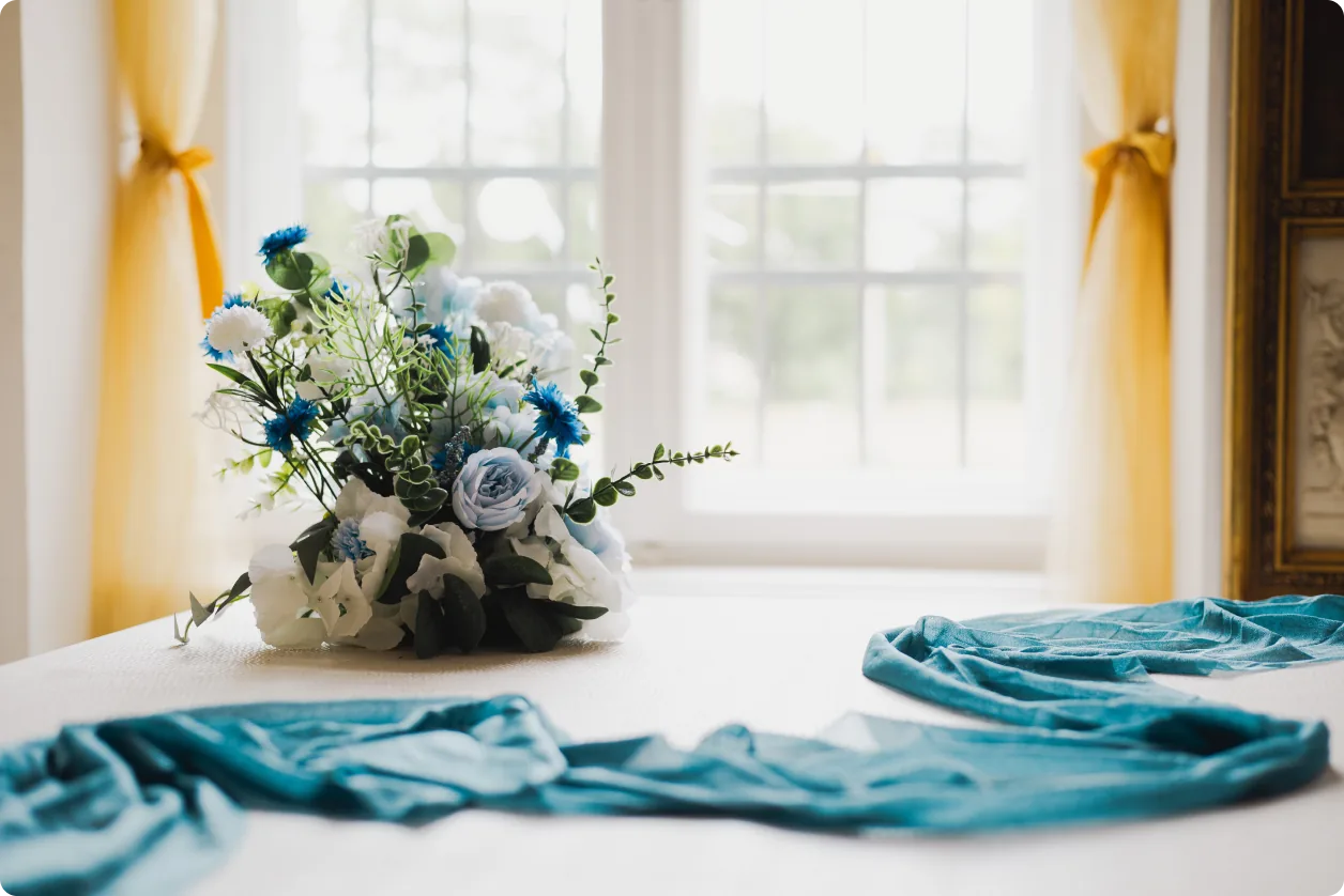 Ein Blumengesteck mit blauen und weißen Blüten steht auf einem Tisch vor einem Fenster mit gelben Vorhängen; davor liegt ein türkisfarbenes Tuch.