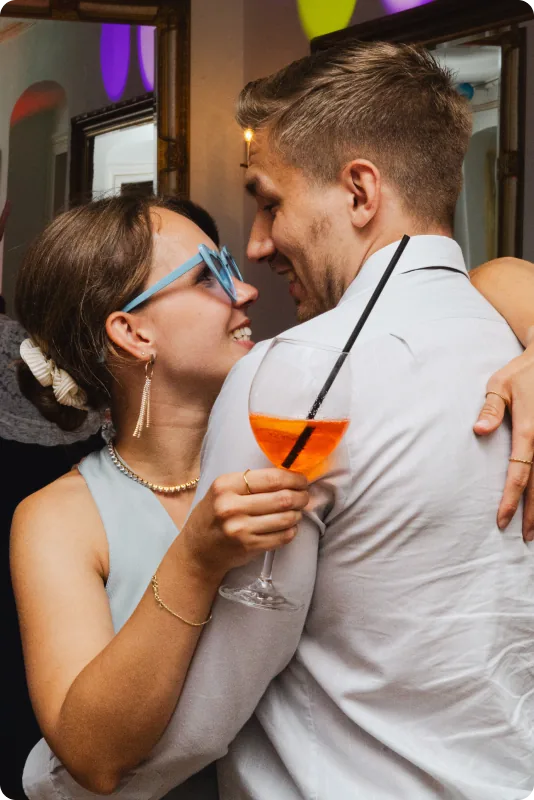 Eine Frau mit blauer Brille und ein Mann im weißen Hemd tanzen eng umschlungen; sie hält ein Glas mit orangefarbenem Getränk.