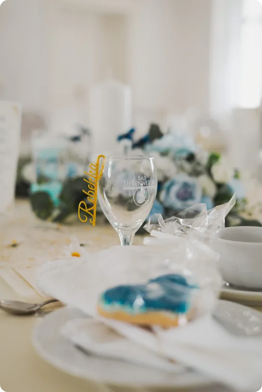 Ein Namensschild aus goldenem Draht mit dem Schriftzug „Rebekka“ steckt an einem Glas auf der Hochzeitstafel; im Vordergrund ein verpacktes Herzgebäck, dahinter verschwimmen blaue Blumen und Kerzen.