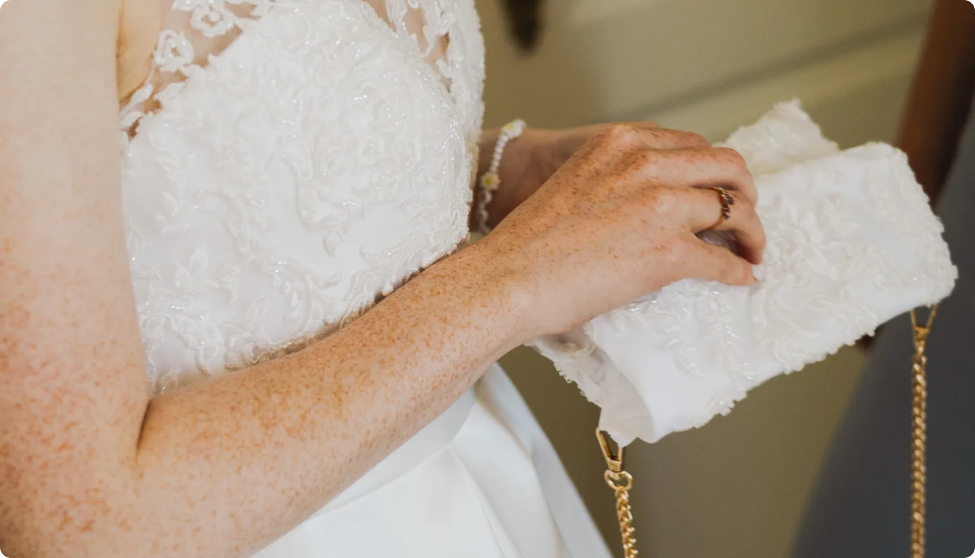 Nahaufnahme der Hand der Braut mit Ring, die eine mit Spitze besetzte weiße Clutch hält; ihr Kleid ist mit feiner Spitze verziert.