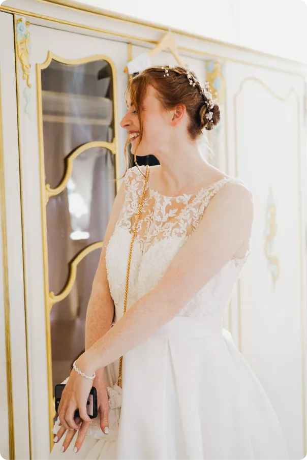 Die Braut mit Haarschmuck und Spitzenkleid lacht und blickt zur Seite; sie steht vor einem hellen Schrank mit goldenen Verzierungen und hält ihr Handy.
