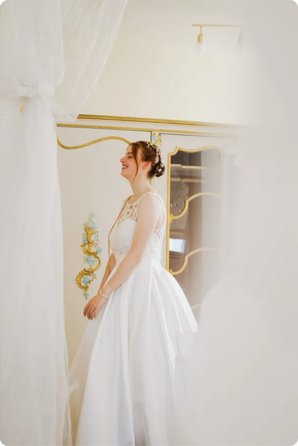 Die Braut im weißen Kleid steht lachend zwischen einem durchsichtigen Vorhang und einem Schrank mit goldenen Verzierungen.