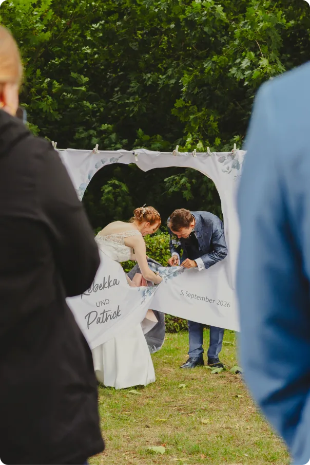 Braut und Bräutigam schneiden gemeinsam ein Herz aus einem gespannten Laken mit der Aufschrift „Rebekka und Patrick“ und dem Datum 5. September 2026.