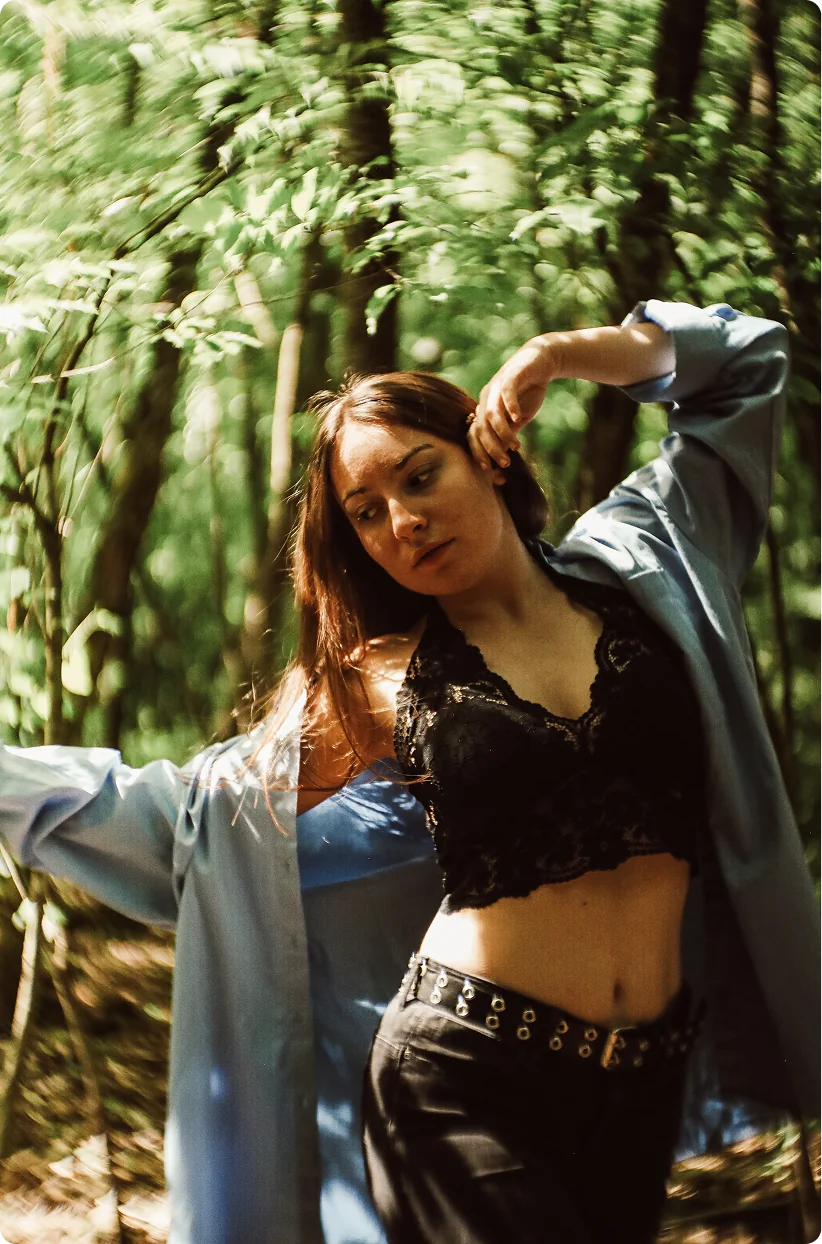 Eine Frau in offenem, hellblauem Hemd und schwarzem Spitzentop hebt einen Arm hinter den Kopf und blickt zur Seite; sie steht in einem sonnigen Wald.