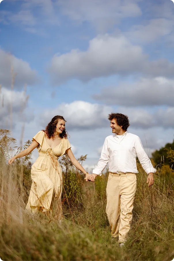 Ein lachendes Paar läuft Hand in Hand durch hohes Gras unter blauem Wolkenhimmel; sie trägt ein gelbes Kleid, er ein weißes Hemd und beige Hosen.
