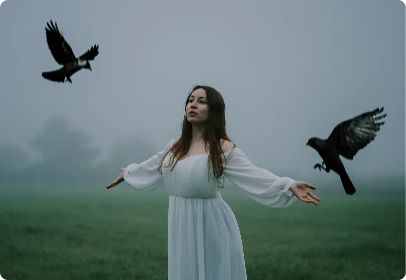 Eine Frau in einem weißen Kleid steht mit ausgebreiteten Armen auf einer nebligen Wiese, links und rechts von ihr fliegen schwarze Raben.