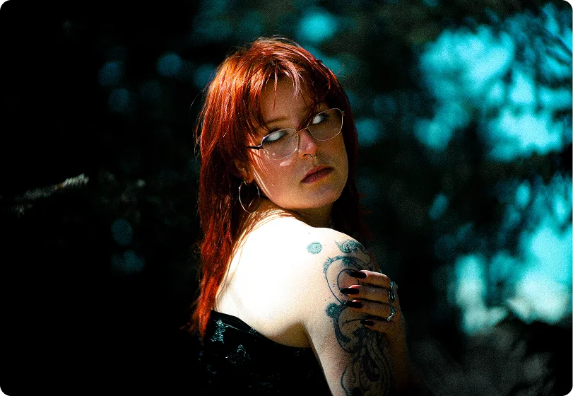 Eine Frau mit rotem Haar und Brille blickt über ihre tätowierte Schulter in die Kamera; hinter ihr schimmert dunkler Wald mit türkisfarbenen Lichtreflexen.