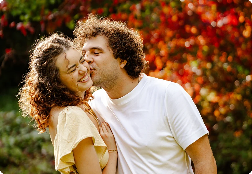 Eine Frau mit lockigem Haar lacht herzlich, während ein Mann mit dunklen Locken ihr einen Kuss auf die Wange gibt; dahinter leuchtet rotes und oranges Herbstlaub.