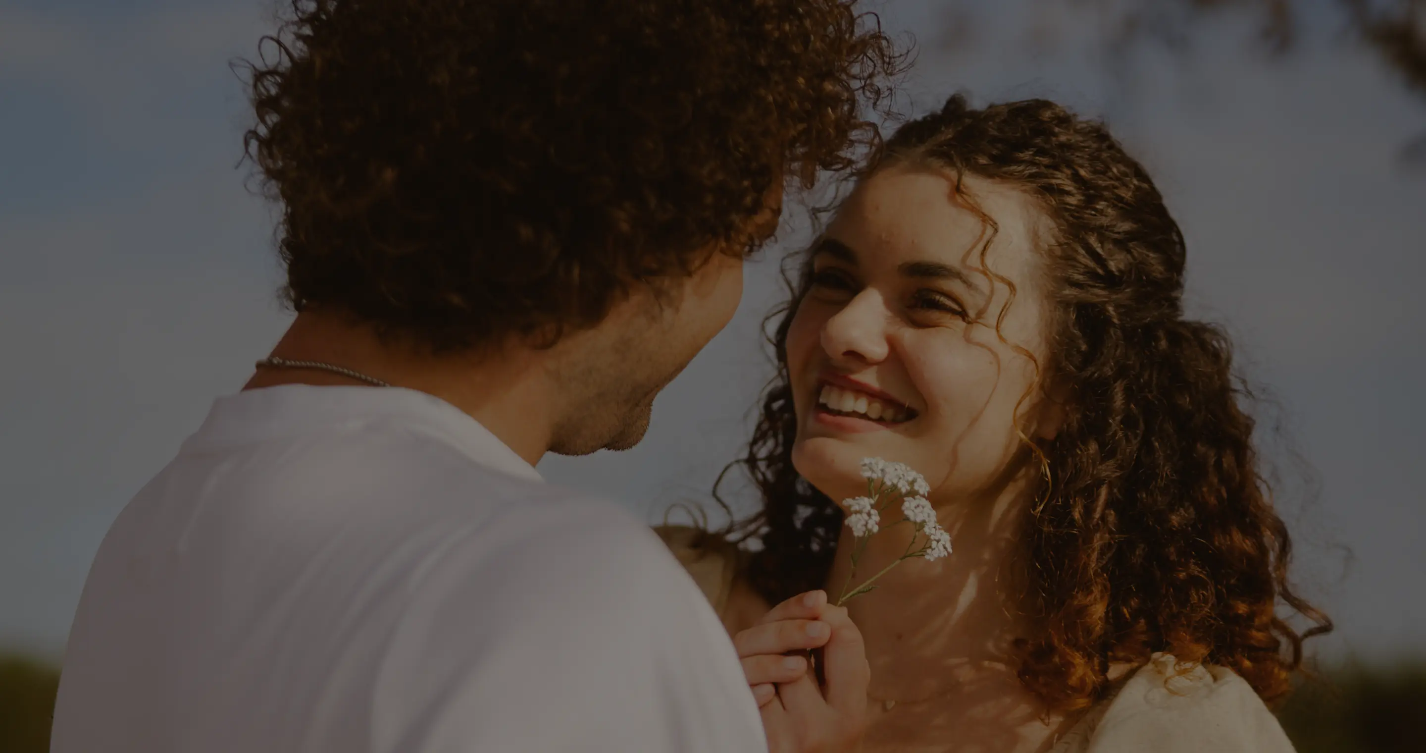 Ein Mann mit dunklen Locken, von hinten zu sehen, steht vor einer lachenden Frau mit lockigem Haar, die einen kleinen weißen Blumenzweig in den Händen hält.