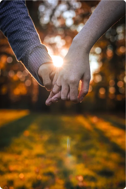 Zwei ineinander gelegte Hände im Gegenlicht, die untergehende Sonne funkelt zwischen den Fingern; im Hintergrund leuchten goldene Herbstbäume.