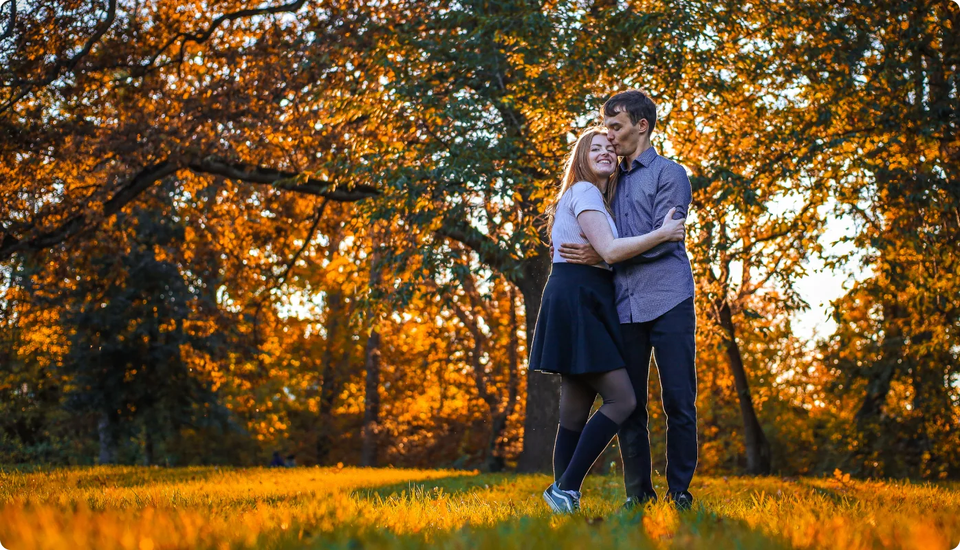 Ein Paar steht umarmt auf einer goldenen Wiese unter orange gefärbten Herbstbäumen; er küsst sie auf die Stirn, sie lächelt.