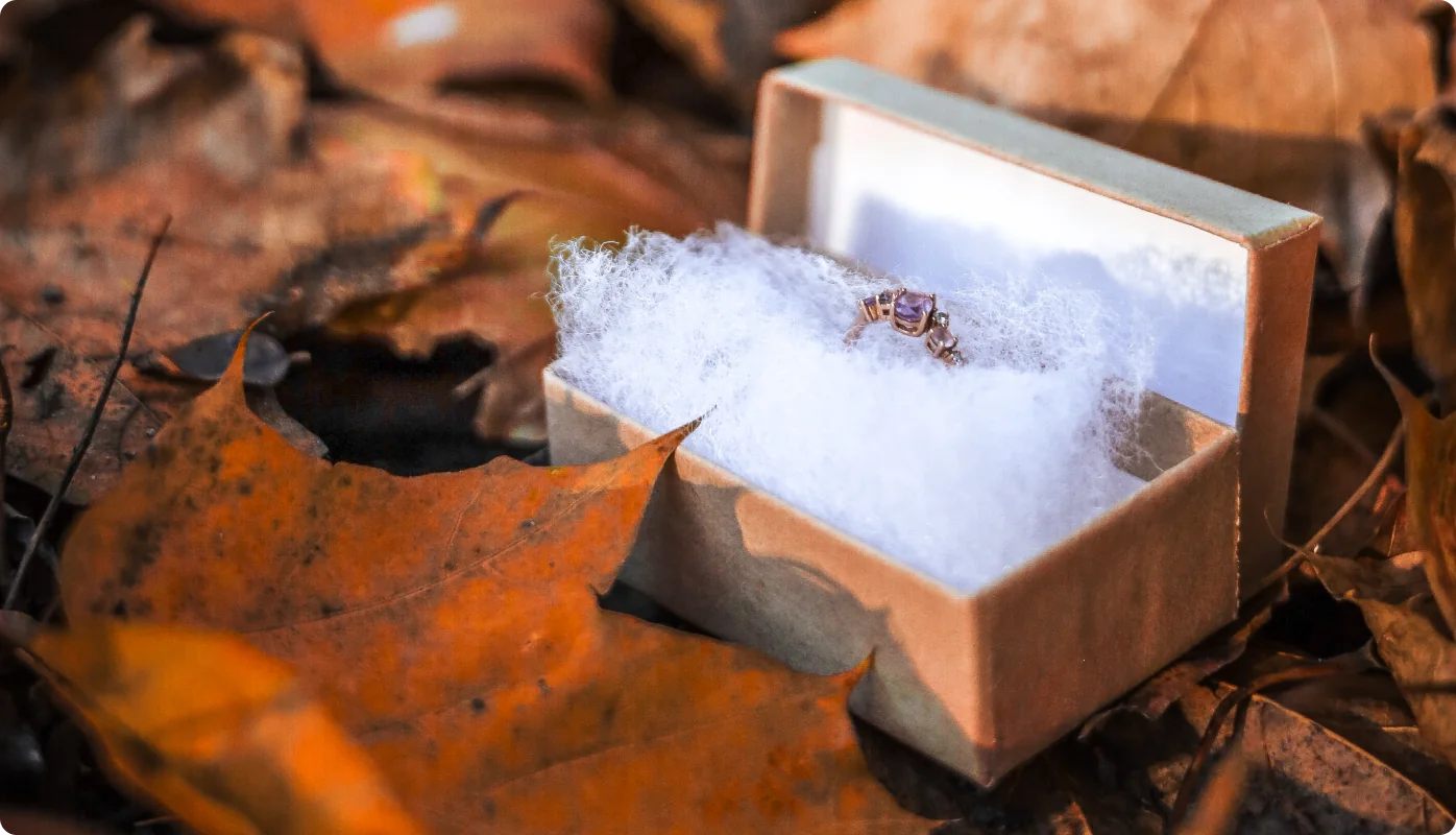 Ein Ring mit violettem Stein liegt in einer geöffneten Schachtel auf weißer Watte, umgeben von orangefarbenen Herbstblättern.